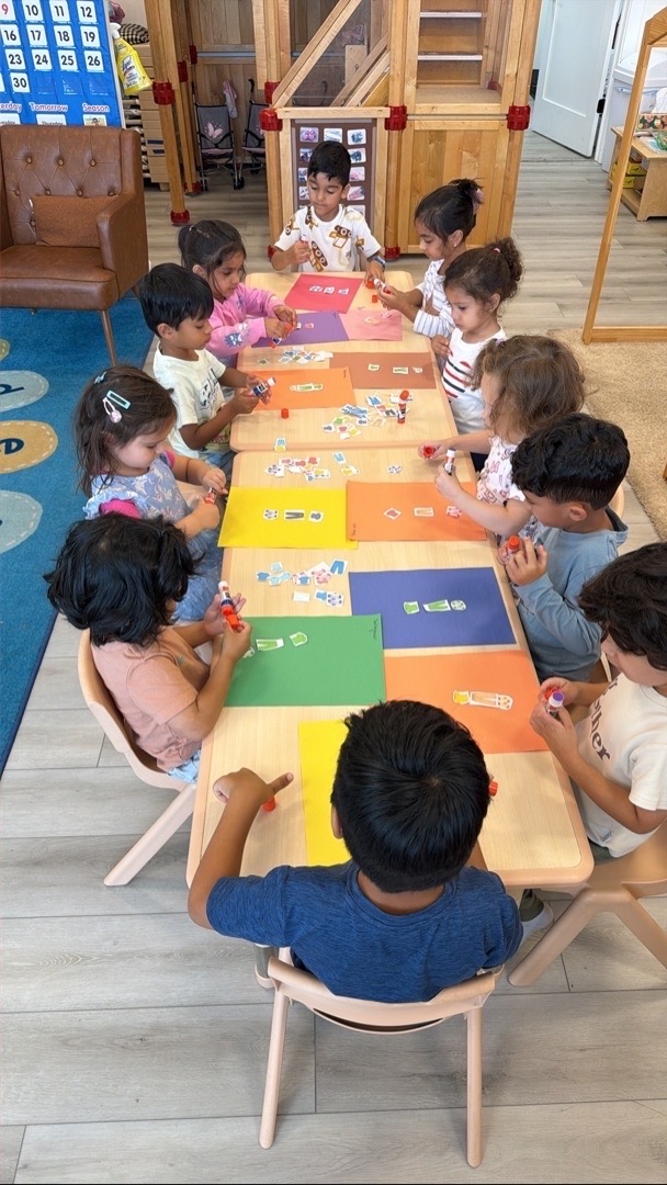 Children gluing paper shapes onto colored sheets at a classroom table
